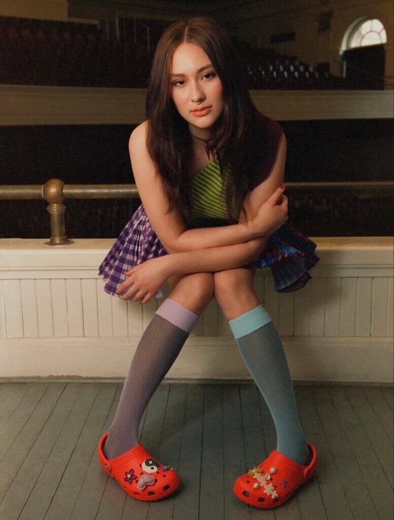 Lola Tung poses in a green top, purple and black gingham skirt, knee-high socks and bright red Crocs in an empty theater.