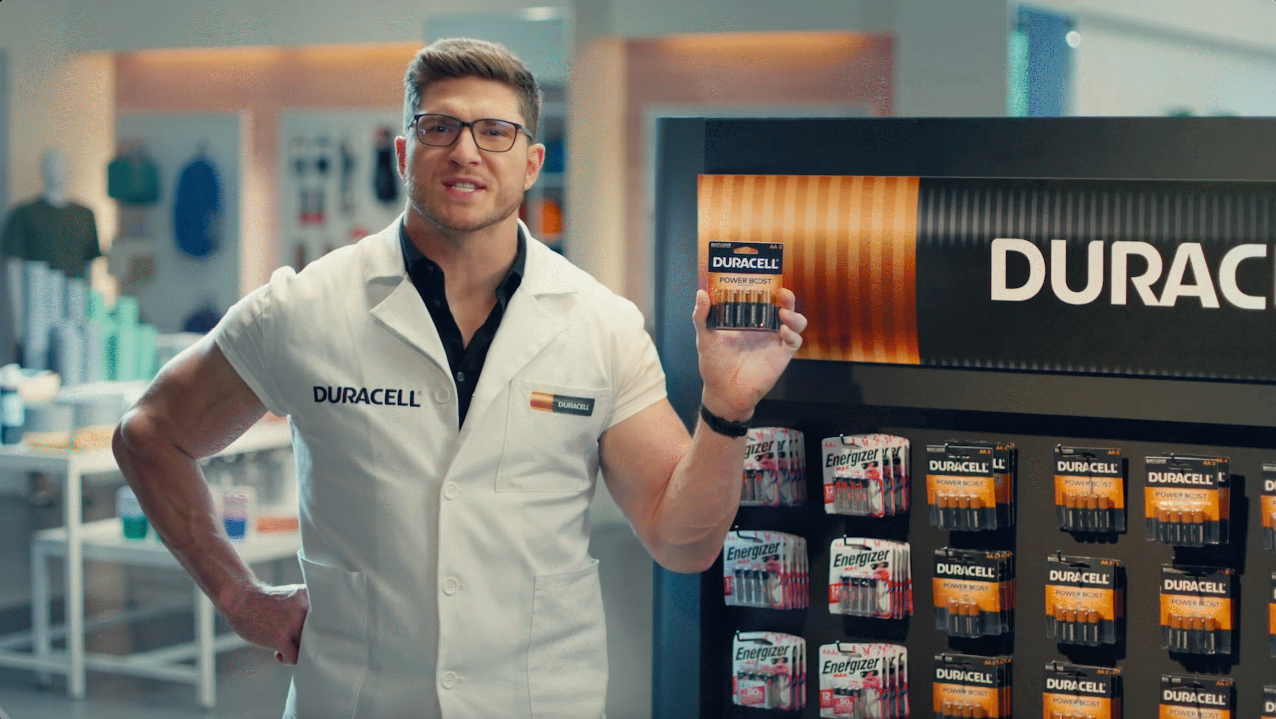 A buff white man in a Duracell lab coat stands in a store by a Duracell battery display and proudly holds up a pack of batteries.
