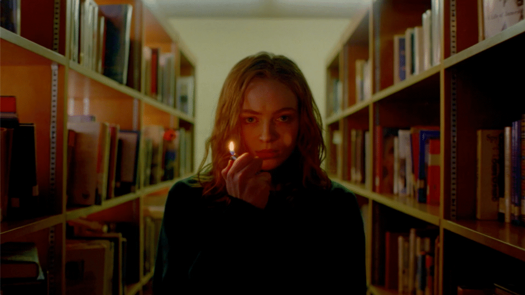 Sadie Sink stands in a dimly lit library, illuminating her face with a lighter.