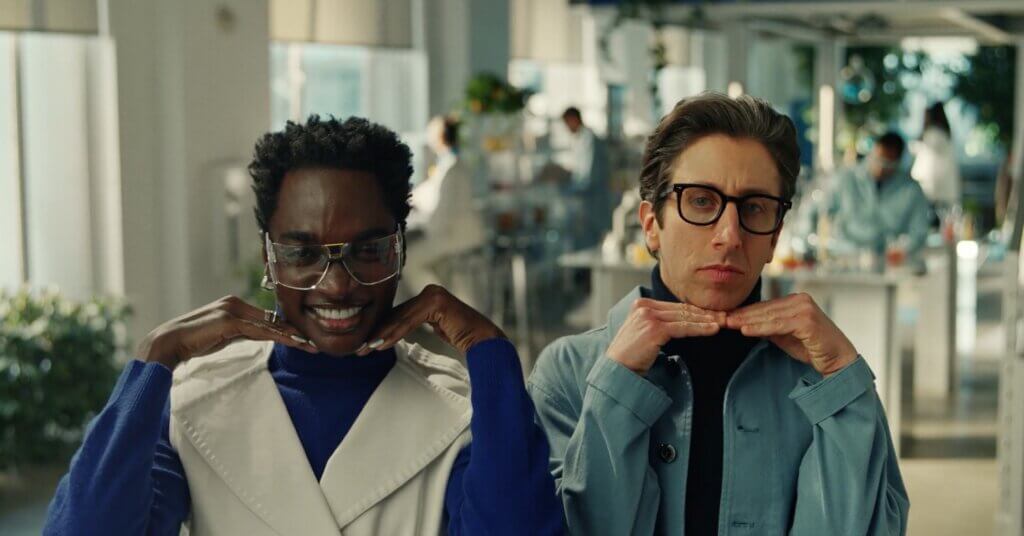 Rickey Thompson and Simon Helberg pose. resting their chins on their bent hands. Behind them is a brightly-lit lab filled with greenery.