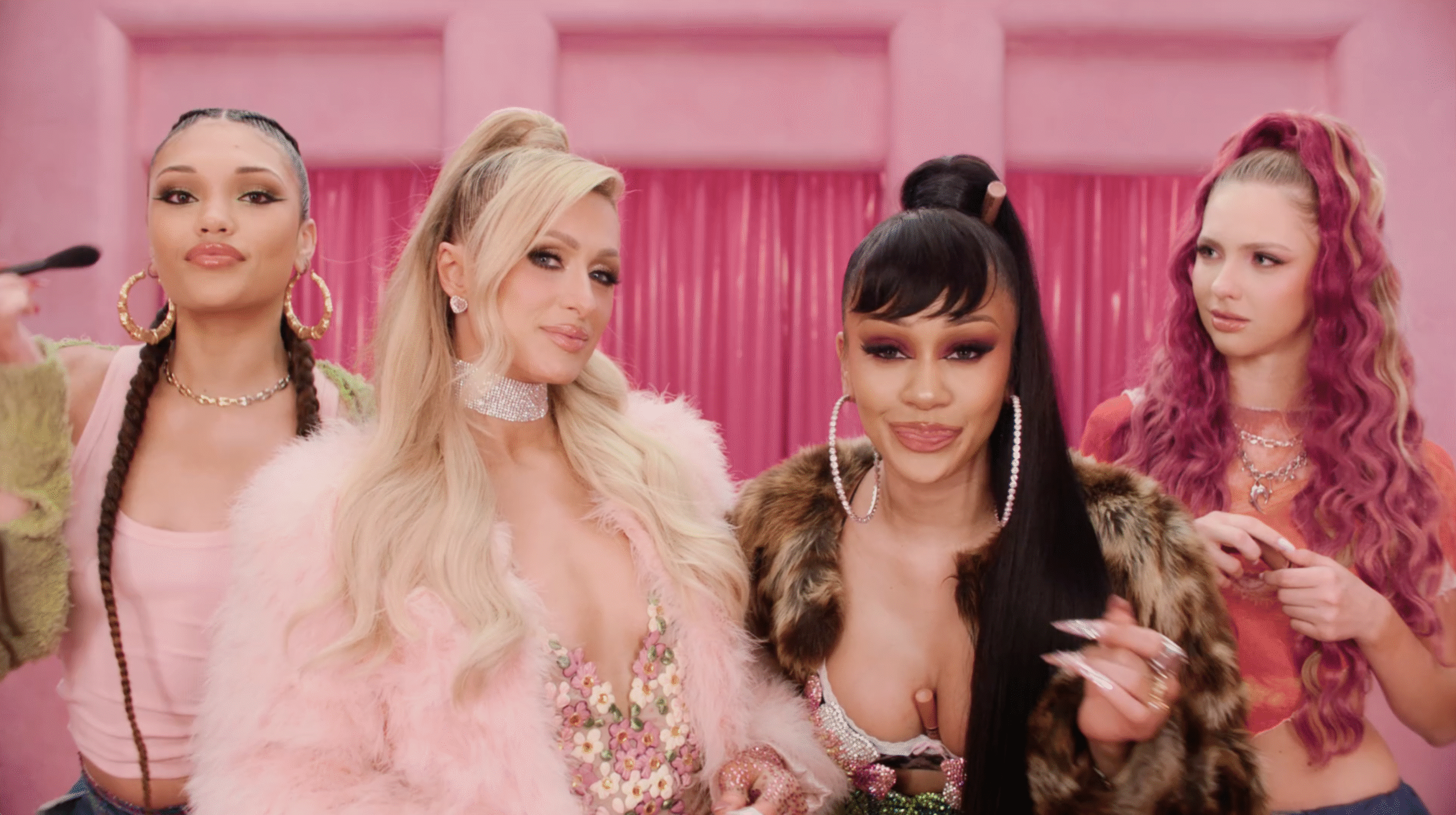 Paris Hilton and Saweetie stand between two teenagers, one with two braids and the other with pink curly hair. They are in a brightly-lit pink room and are leaning into the camera.