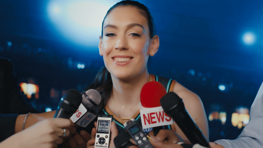 Breanna Stewart stands in front of a blue flashing screen smiling at the camera as different kinds of microphones are held up to her.
