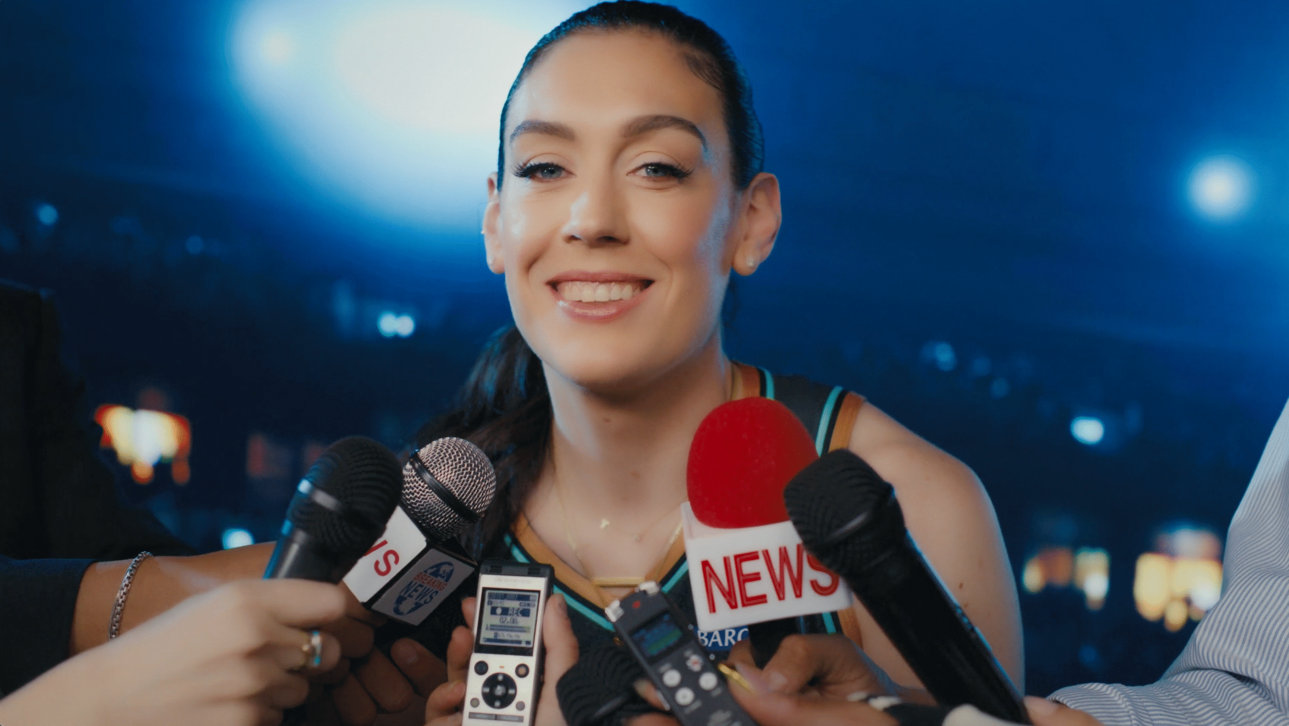Breanna Stewart stands in front of a blue flashing screen smiling at the camera as different kinds of microphones are held up to her.
