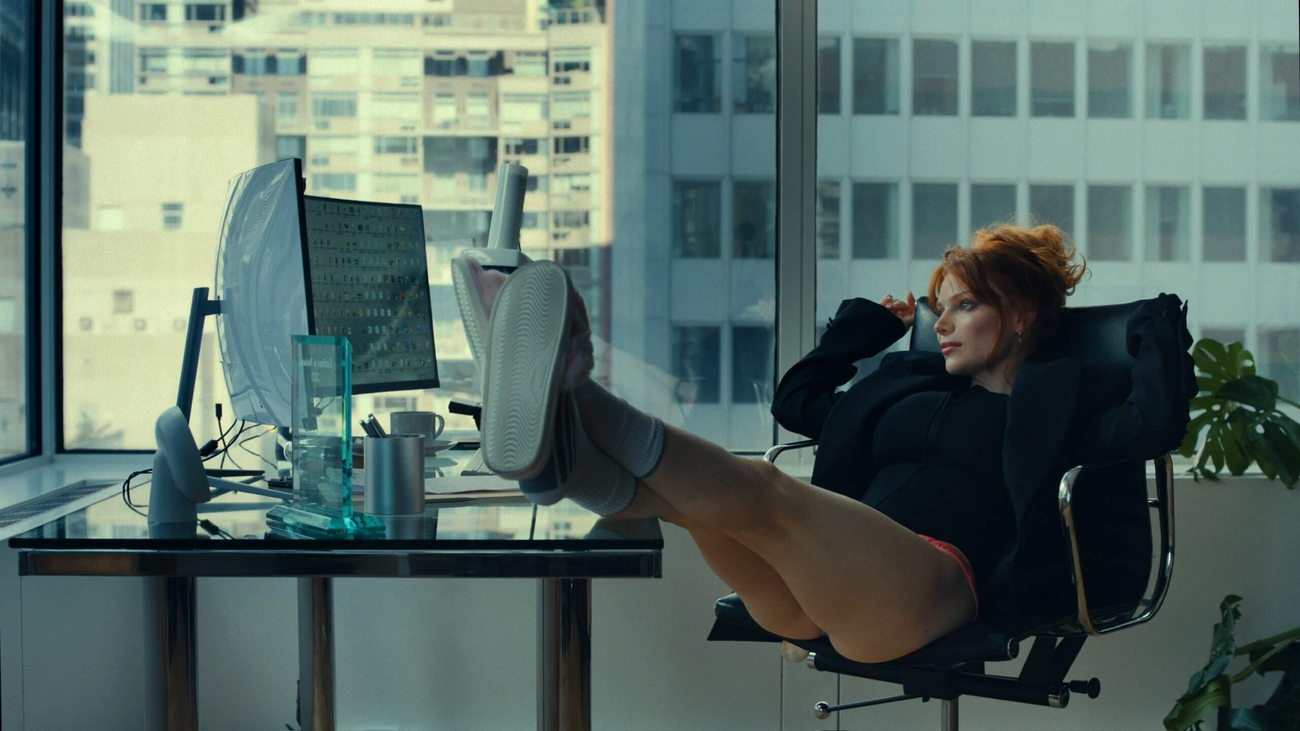 Levi Coralynn reclines in an office chair, putting her feet on the desk. She has two monitors in front of her and sits in front of large windows showing a building behind her.