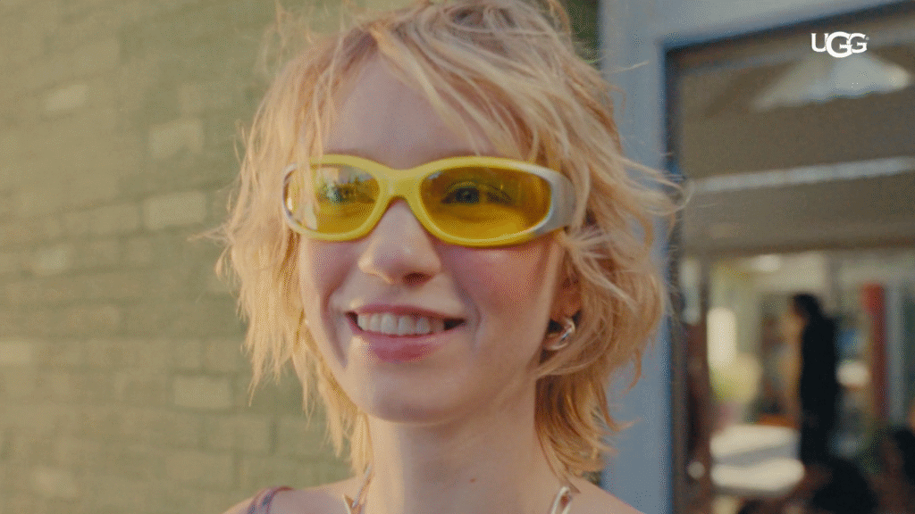 A blonde white girl with short hair smiles outside of a house on a bright sunny day. She is wearing big futuristic yellow sunglasses.